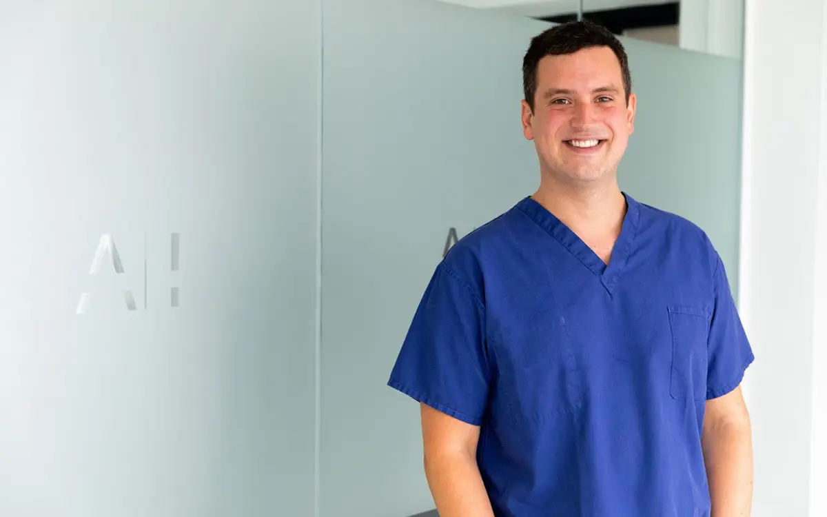 A smiling individual wearing blue medical scrubs stands in front of a frosted glass panel at Healthium Clinics. The blurred letters AH are visible on the left side of the panel, hinting at their location in Henley-On-Thames.