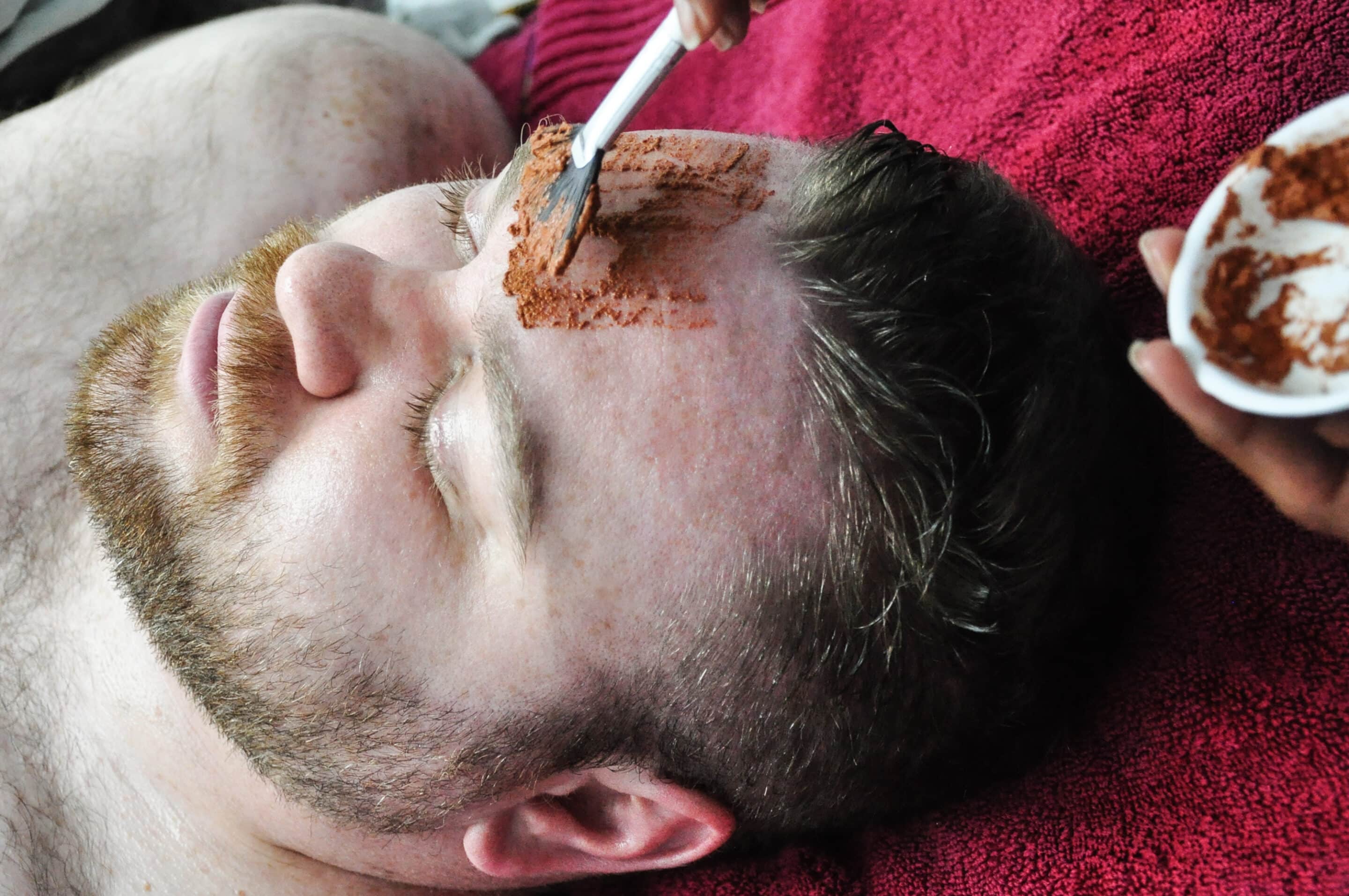 A bearded man is lying down with his eyes closed while a hand applies a brown facial mask to his forehead using a brush. A small bowl with more of the mask is visible in the other hand. Hes resting on a red towel.