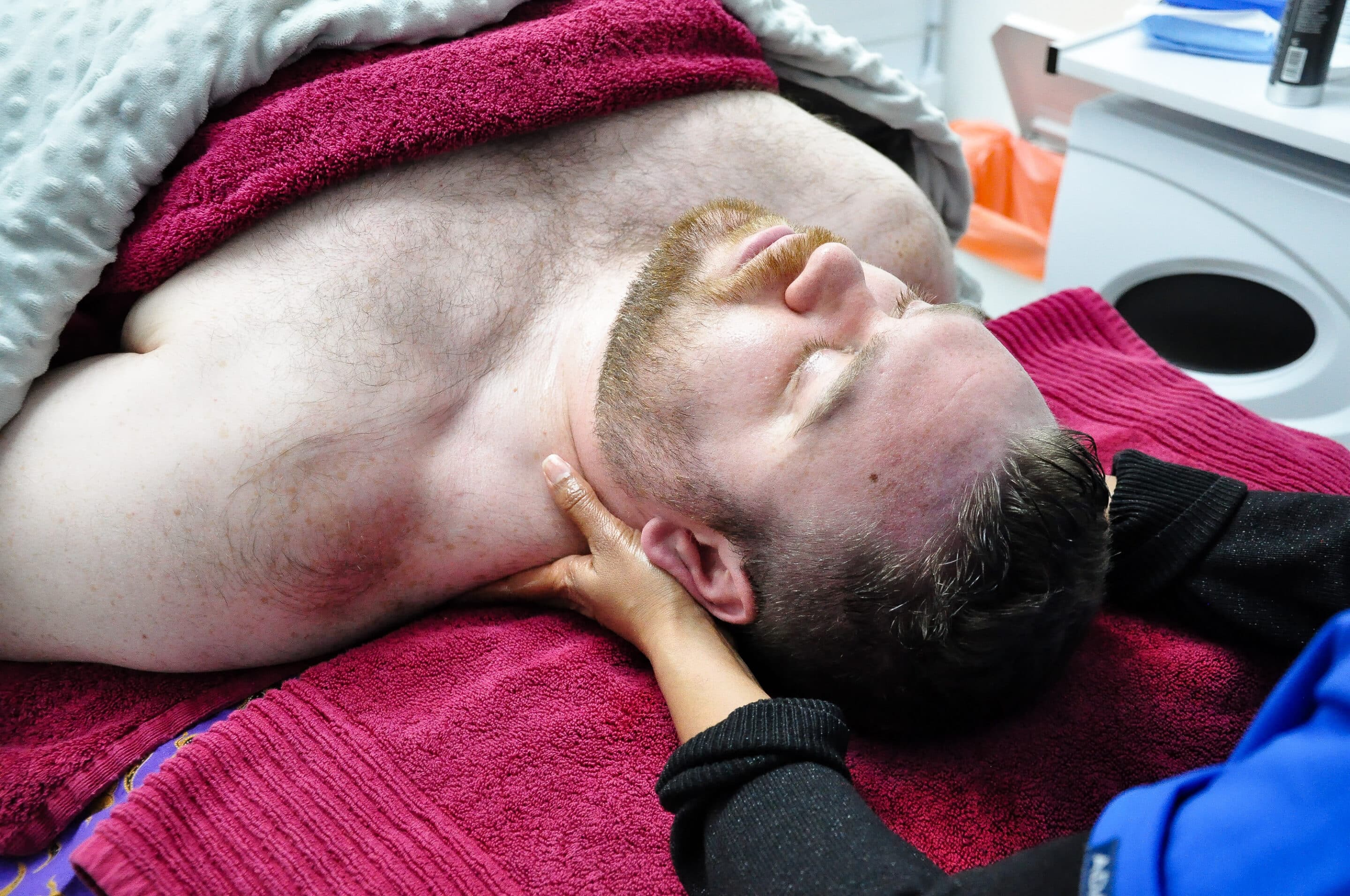 A man with a beard lies on a massage table, eyes closed, covered with red and gray towels. A person is using their hands to massage the mans neck and shoulders. Nearby, a white cabinet and some small items are visible.