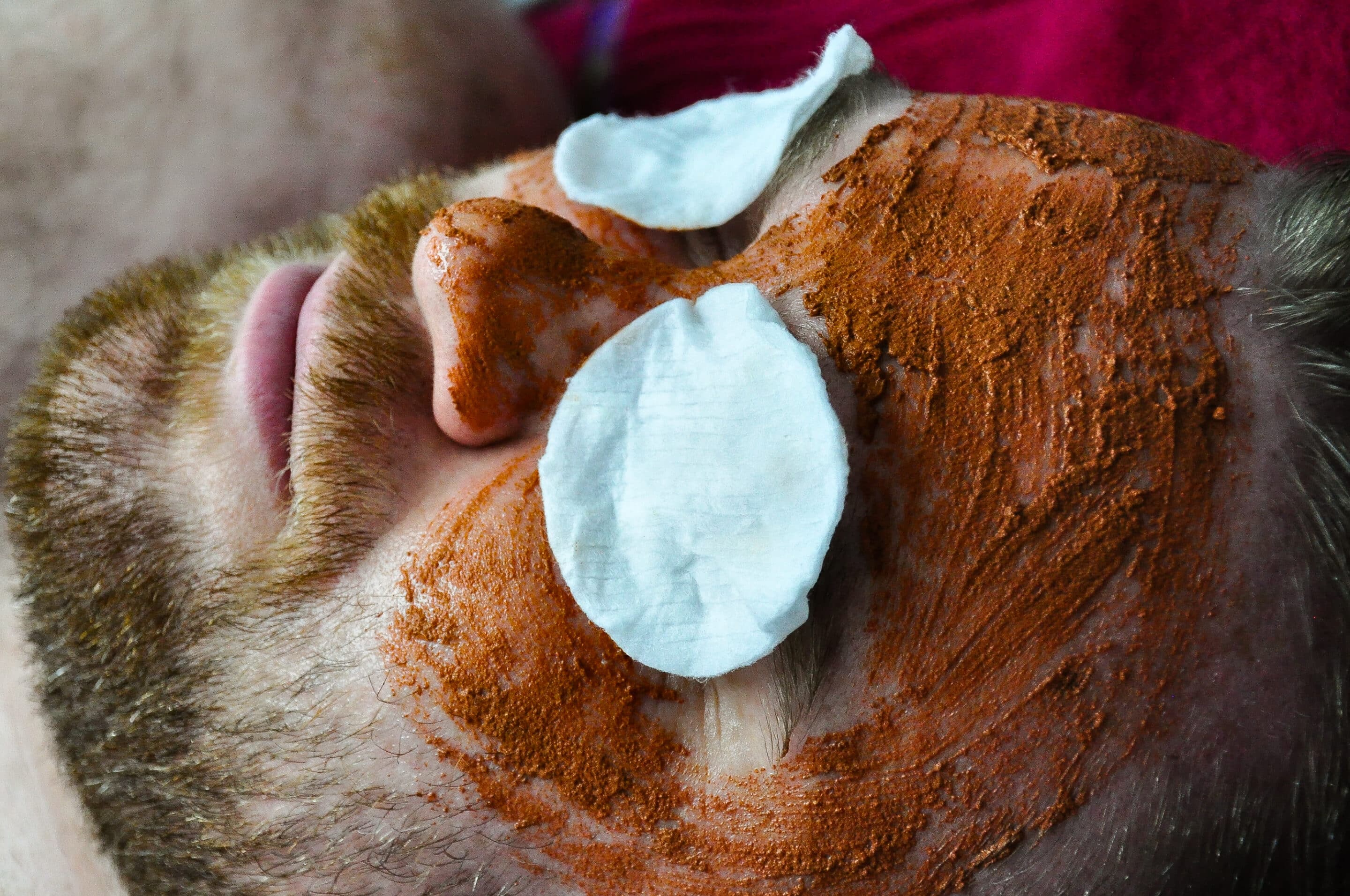 Beauty-facial A person with a red beard is lying down with a reddish-brown facial mask applied to their face. Two cotton pads are placed over their eyes.