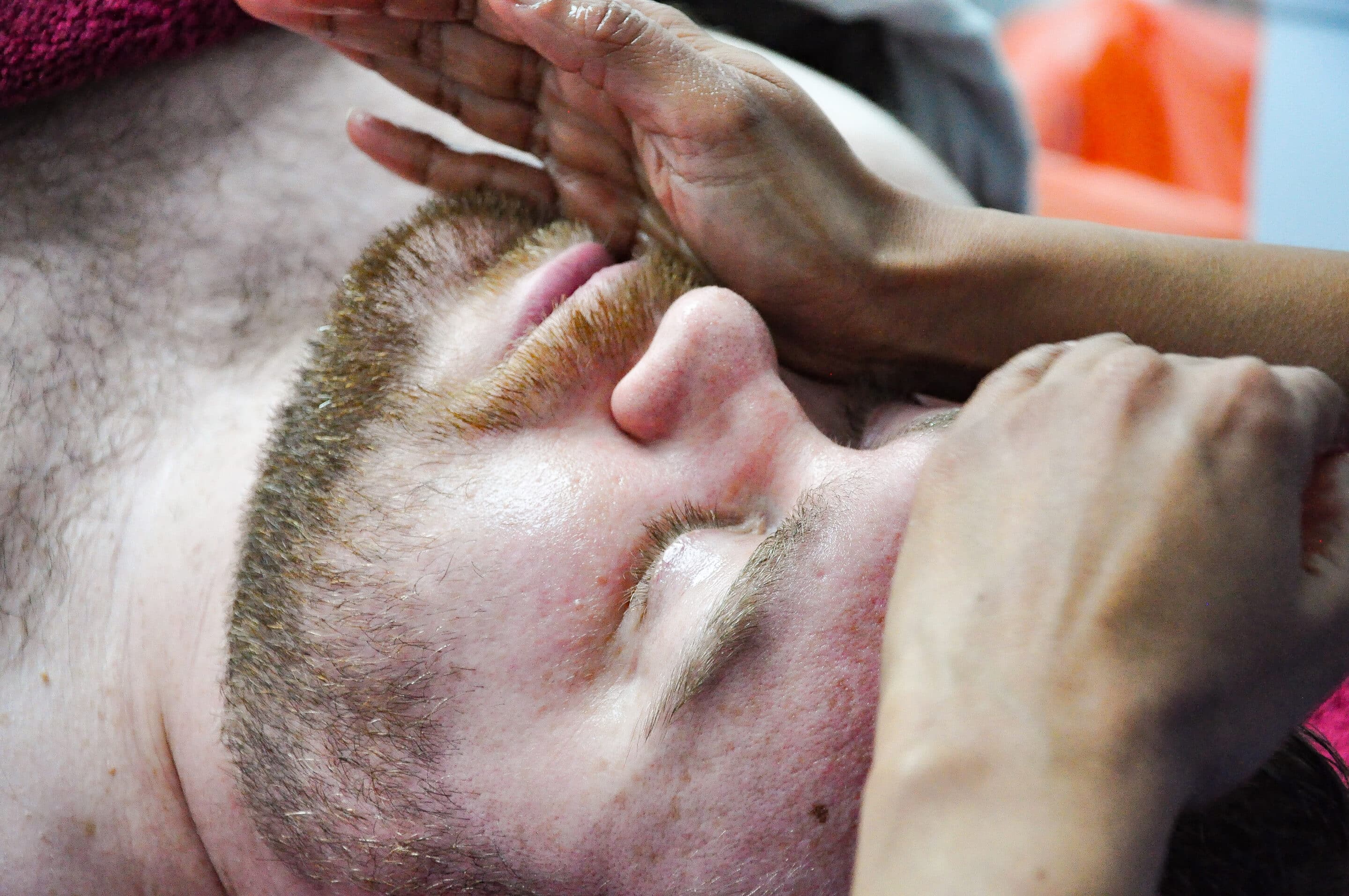 facial-massage A person with a beard is lying down with eyes closed, receiving a facial massage from another persons hands. The bearded individual is covered with a towel, and the background is blurred.