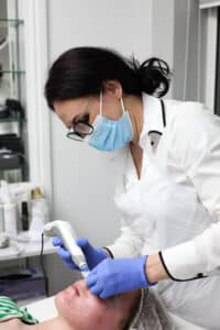 A woman in a white coat, wearing a blue mask and purple gloves, uses a skincare device on another womans face in a clinic. The patient is lying down, covered with a hair cap. Skincare products are visible in the background.