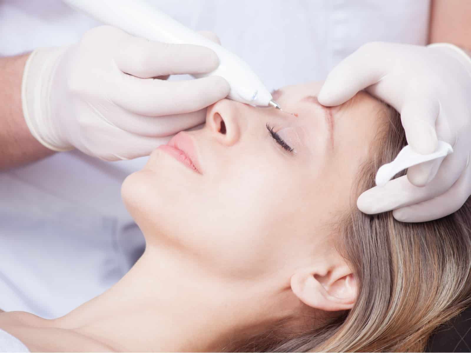 A person is lying down with their eyes closed as a professional, wearing white gloves, uses a cosmetic device on their eyelid. The setting appears to be a beauty or medical spa.