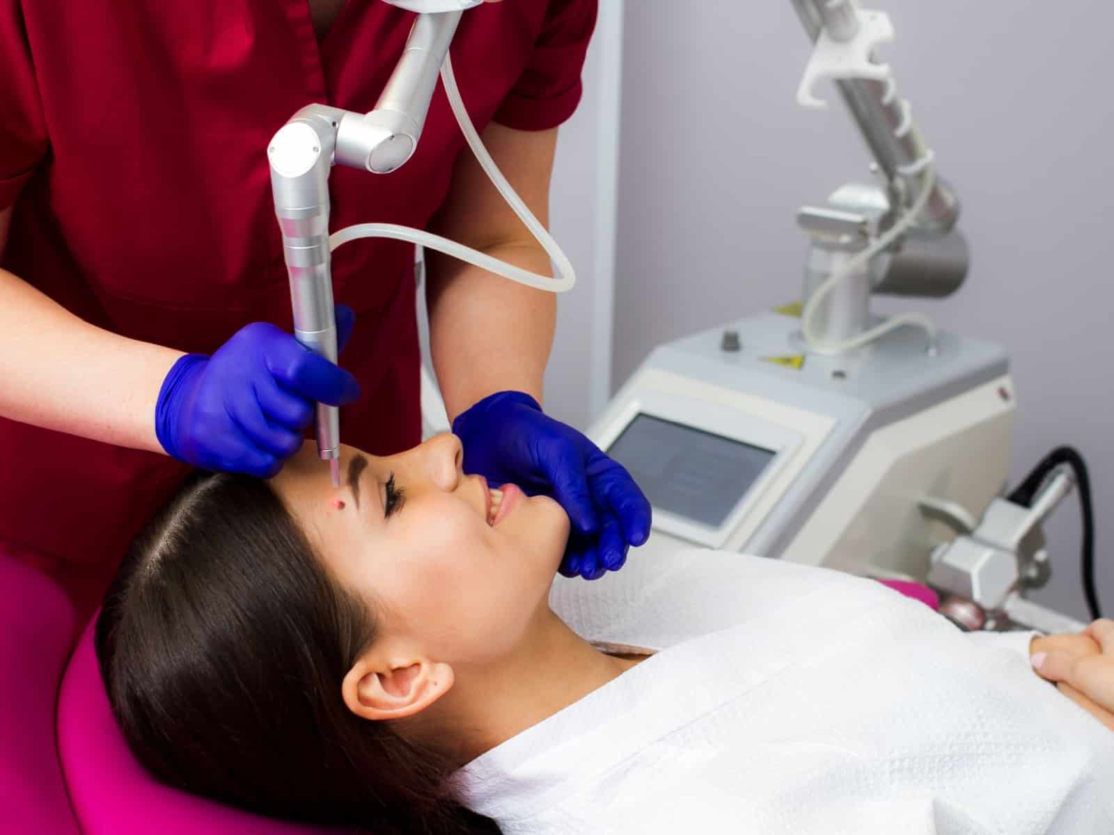 Untitled A woman lying on a chair receives a laser treatment on her face. A professional wearing purple gloves operates the laser device. The woman is wearing a white gown and appears relaxed. Nearby is a machine with a digital display.