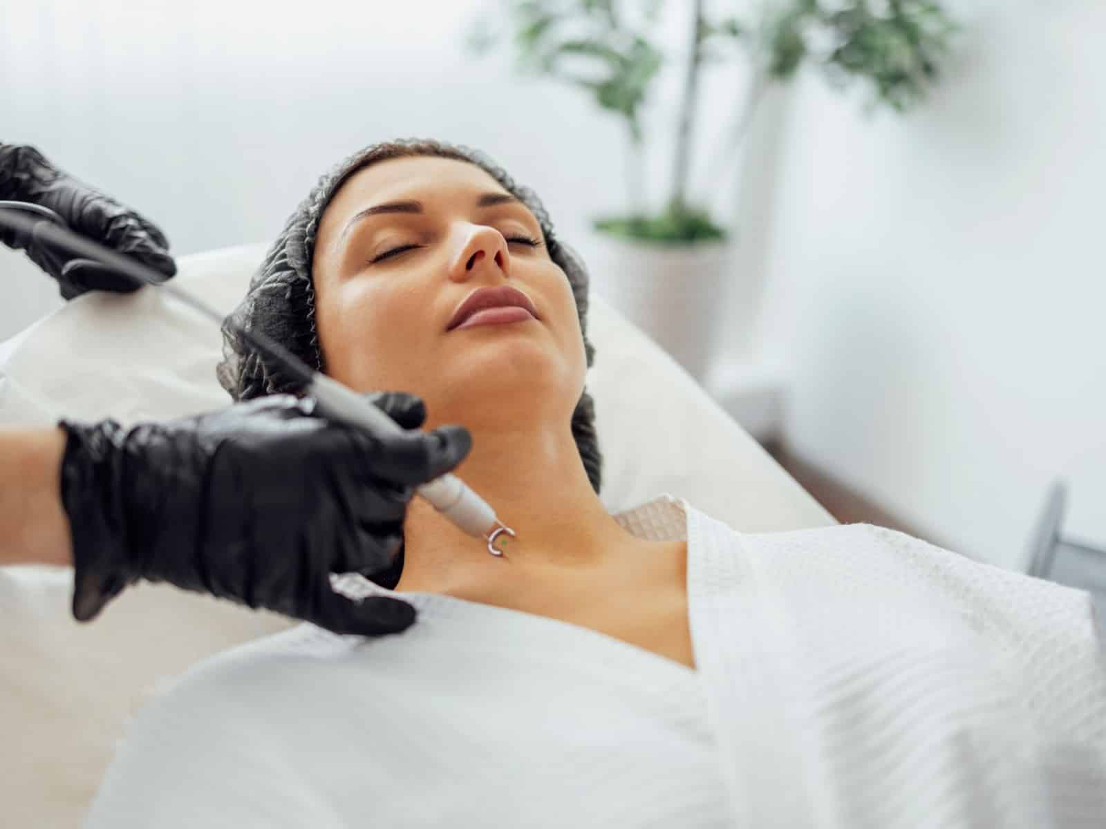 A woman with her eyes closed is receiving a cosmetic procedure on her face. She is wearing a white robe and a protective cap, while a gloved technician holds a device to her neck. A potted plant is in the softly focused background.