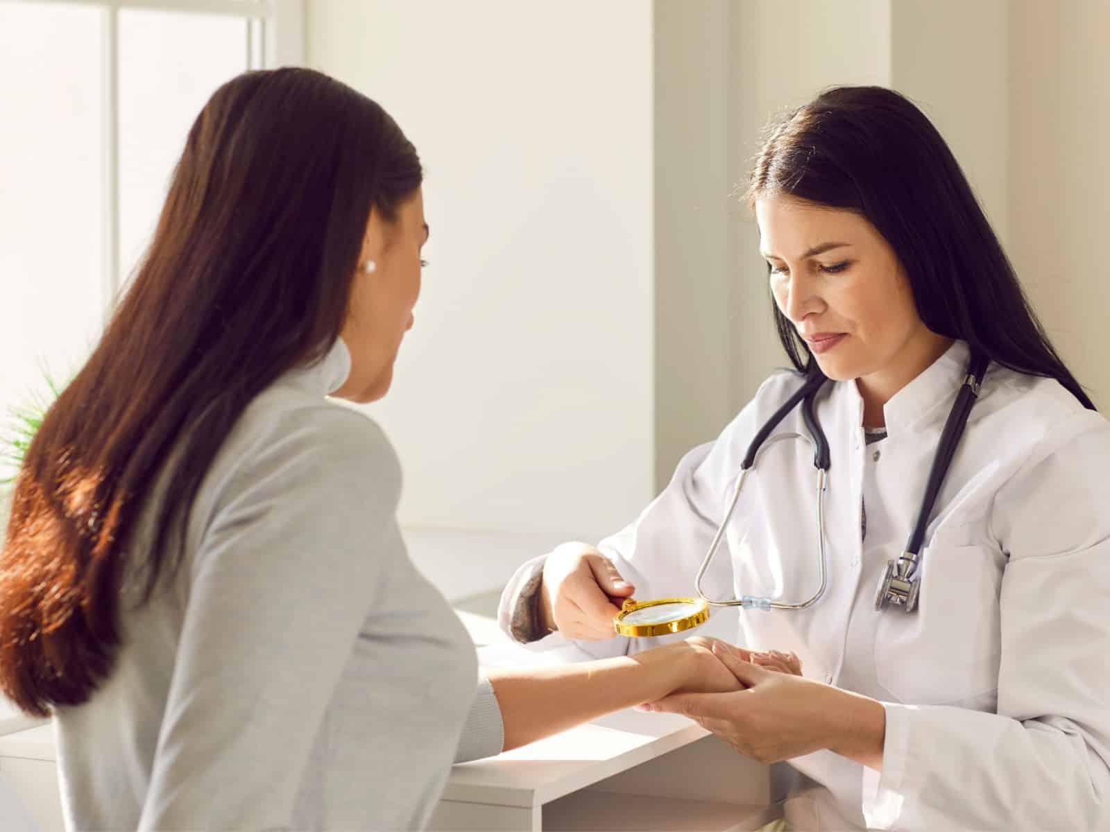 Untitled A doctor in a white coat examines a patients hand with a magnifying glass at a desk. The patient, with long brown hair, sits across from the doctor in a bright, sunlit room. Both are focused on the examination.