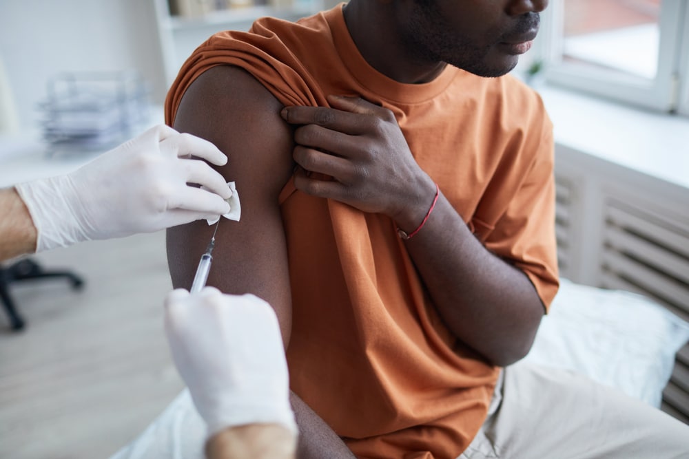 Close,Up,Of,Adult,African-american,Man,Looking,Away,While,Getting A person in an orange shirt sits in a clinic, receiving a vaccination. They hold up their sleeve as a healthcare professional in white gloves administers the shot using a syringe. The setting is bright, with a window in the background.