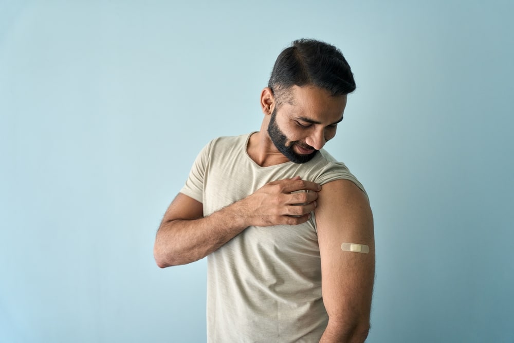 Happy,Healthy,Young,Indian,Man,Showing,Bandage,Plaster,On,Arm A man with a beard, wearing a beige t-shirt, looks at a bandage on his upper arm against a light blue background.