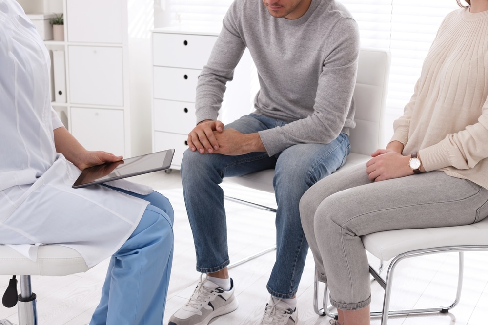 Couple,Having,Appointment,With,Std,Specialist,In,Clinic,,Closeup A healthcare professional sits with a tablet, talking to a seated couple. The man in a gray sweater and jeans rests his hands together, while the woman in a beige sweater has her hands on her lap. The setting is a bright, modern office.