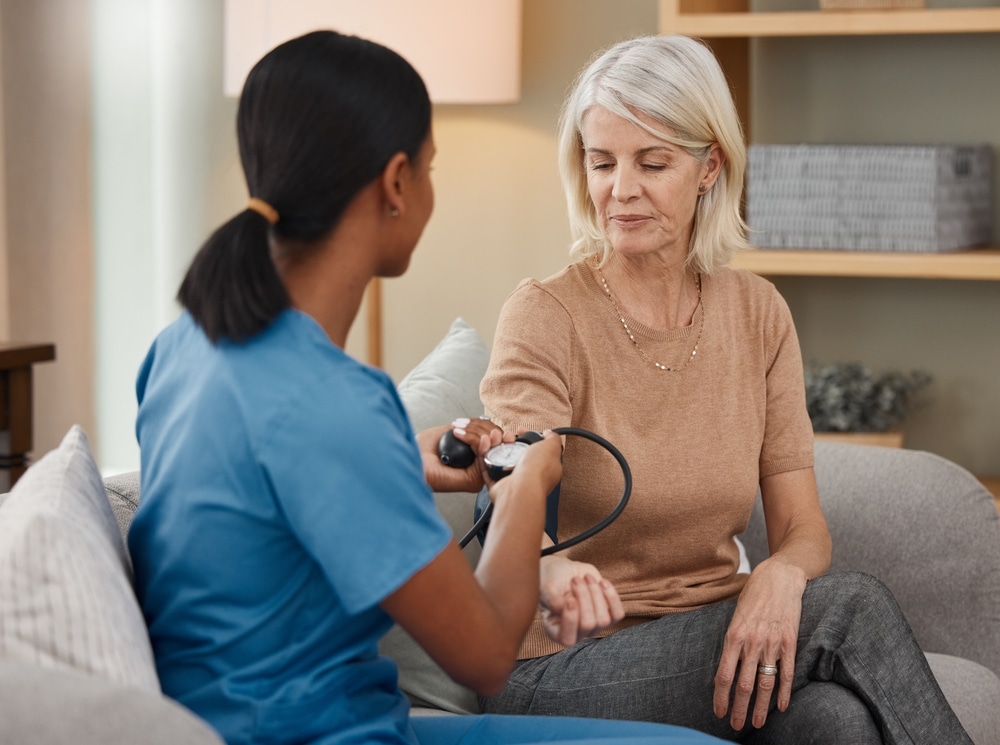 Lets,Hope,My,Hypertension,Is,Under,Control.,Shot,Of,A A healthcare professional in blue scrubs is taking the blood pressure of a woman sitting on a sofa. The woman is wearing a brown top and has short blonde hair. The setting is cozy, with a lamp and bookshelf in the background.