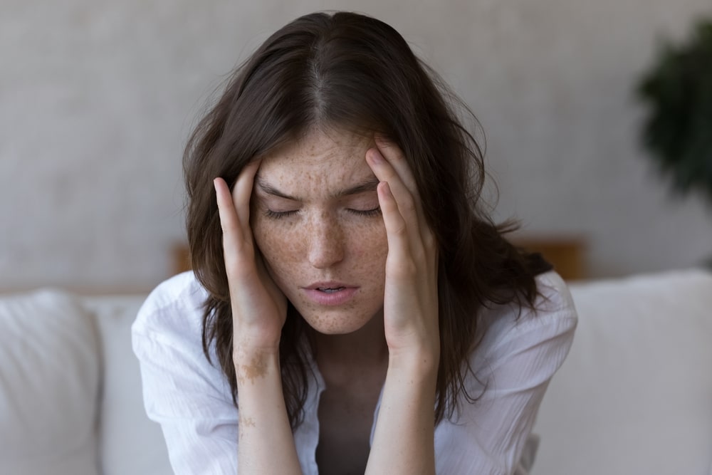 Young,Freckled,Woman,Closed,Eyes,,Touches,Her,Temples,Suffers,From A woman with long brown hair, wearing a white shirt, sits on a couch with her eyes closed. She rests her hands on her temples, appearing stressed or in pain. The background is softly blurred.