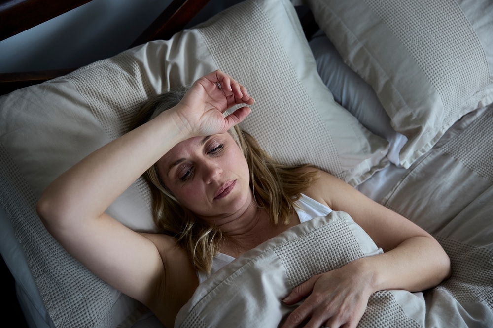 Menopausal,Mature,Woman,Suffering,With,Insomnia,In,Bed,At,Home A woman lies in bed under a beige duvet, looking tired or unwell. Her right hand rests on her forehead, and she has a thoughtful expression. The setting appears to be a dimly lit bedroom with matching pillows.