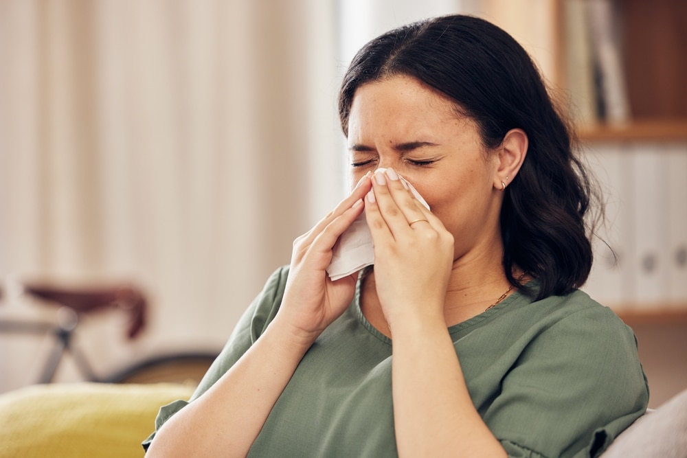 Sick,,Allergy,And,Blowing,Nose,With,Woman,On,Sofa,For A person with dark hair wearing a green shirt is sitting indoors, sneezing into a tissue. Bookshelves and a bicycle are visible in the blurred background.