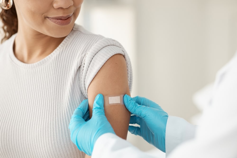 Vaccine,,Happy,Woman,And,Hands,Of,Doctor,With,Plaster,For A person wearing a white top receives a bandage on their upper arm from someone in blue gloves, indicating they have just received an injection or vaccination. The background is softly blurred.