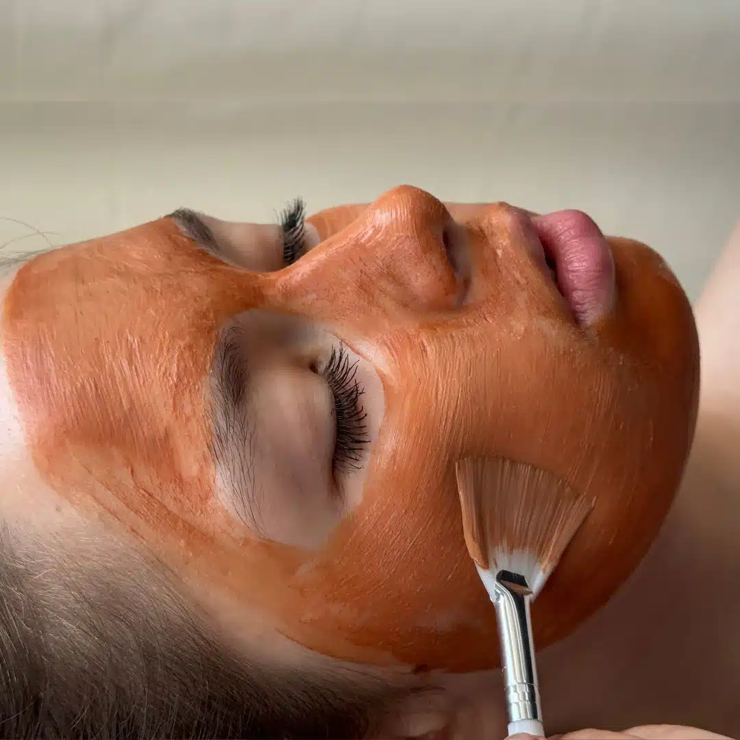 A person lies with eyes closed while a thick orange facial mask is being applied to their face using a soft brush.