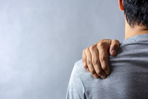 Middle,Aged,Man,Has,Shoulder,Pain.,With,Copy,Space,For A person wearing a gray shirt is shown from behind, holding their left shoulder with their right hand, suggesting pain or discomfort in the shoulder area.