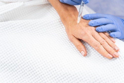 Cropped,Photo,Of,A,Patient,In,A,Waffle,Bathrobe,Being A close-up of a person’s hand receiving an injection, with a medical professional wearing blue gloves administering the shot. The hand rests on a white textured fabric.