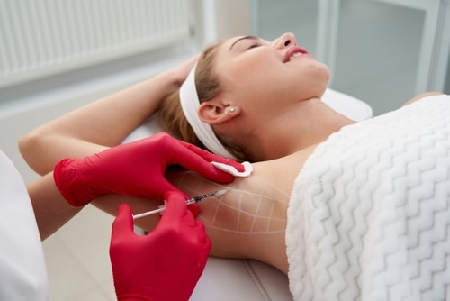 A person lying down with one arm raised, receiving an injection in the underarm area. A practitioner wearing red gloves is administering the injection while holding a cotton pad on the skin.