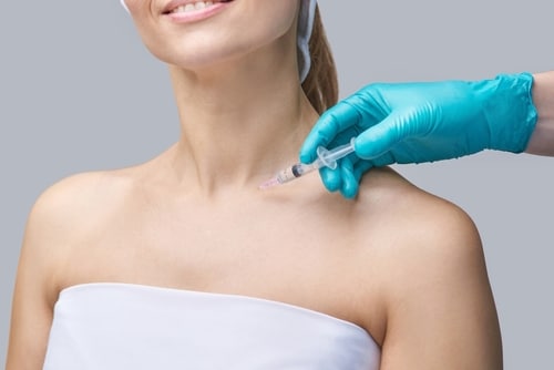 A woman wearing a white towel top and headband receives an injection in her shoulder from a person wearing blue gloves. The background is plain gray. Only the lower face, neck, and shoulders are visible.