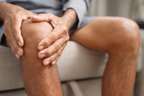 Osteoarthritis,Is,More,Common,In,The,Elderly.,Causes,Knee,Pain, A person sitting on a couch holds their knee with both hands, appearing to experience knee pain or discomfort.