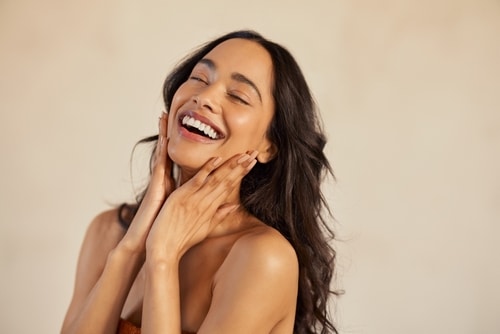 Multiethnic,Young,Woman,Touching,Her,Clean,And,Healthy,Face,Against A woman with long dark hair smiles joyfully with her eyes closed, gently touching her face with both hands. She is wearing a strapless top and standing against a neutral background.