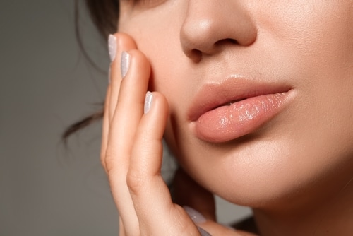 Close-up of a woman gently touching her cheek with her fingers, highlighting smooth skin and natural, glossy lips. Only the lower half of her face is visible.
