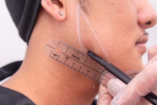 An,Esthetician,Marking,Lines,On,A,Man's,Jaw,Area,As A person with short facial hair has white lines drawn on their jawline. A gloved hand holds a ruler and a pen, marking the jawline for what appears to be a cosmetic or medical procedure.