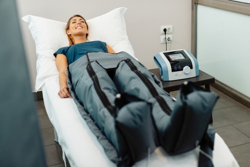Young,Woman,Relaxing,With,Eyes,Closed,During,Pressotherapy,At,Health A woman lies on a treatment bed, smiling, with her legs inside an inflatable compression therapy suit connected to a medical device beside her.