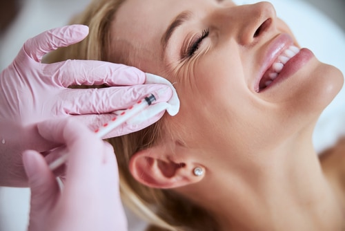 A woman smiling with her eyes closed as a gloved hand administers an injection near her outer eye while holding a cotton pad to her skin.