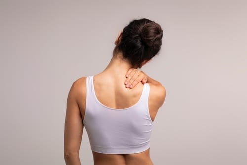 Unrecognizable,Brunette,Woman,With,Neck,And,Back,Pain,Rubbing,Her A woman in a white sports bra faces away, holding her neck with one hand, suggesting neck pain or discomfort. Her hair is tied up in a bun, and the background is plain and light-colored.