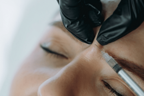 A doctor in a white coat examines a patients hand with a magnifying glass at a desk. The patient, with long brown hair, sits across from the doctor in a bright, sunlit room. Both are focused on the examination.