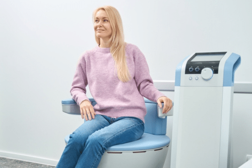 A close-up of a person holding a magnifying glass over a mole on another persons bare back, examining it for medical analysis. The setting appears to be a clinical environment.