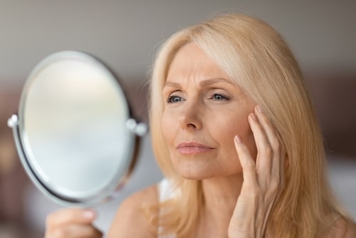 A close-up of a person receiving a laser treatment on a mole near the collarbone. The skin is partially covered with a white towel, and a golden laser device targets the mole.
