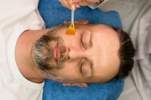 A woman lying on a chair receives a laser treatment on her face. A professional wearing purple gloves operates the laser device. The woman is wearing a white gown and appears relaxed. Nearby is a machine with a digital display.