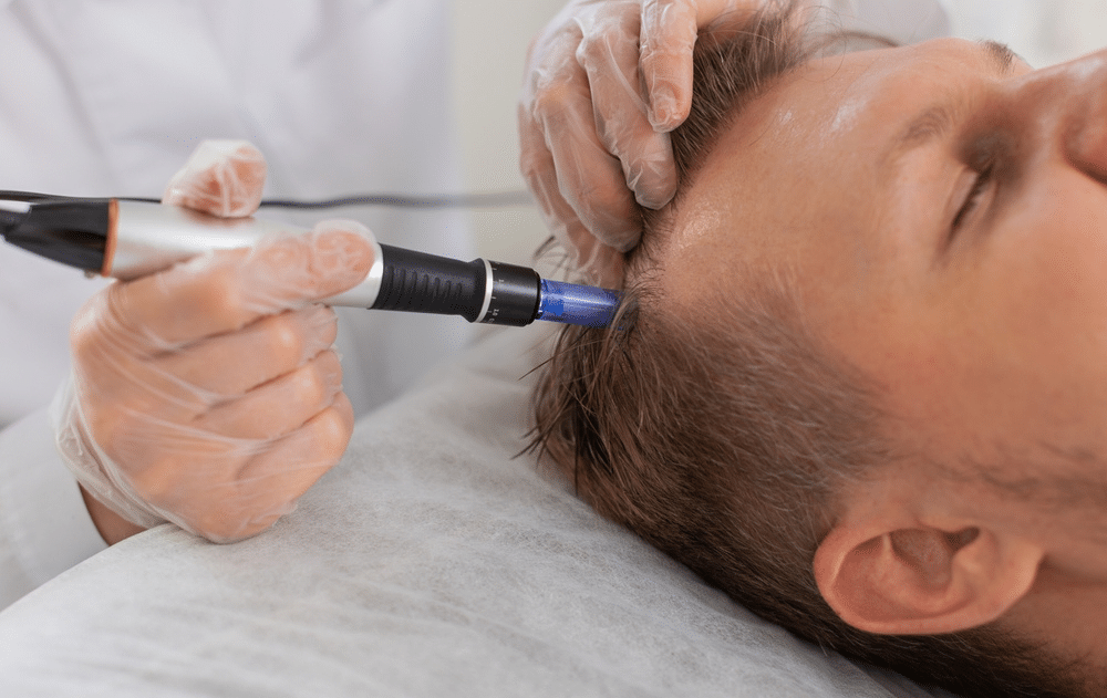 A smiling family of three, with a woman, a man, and a young girl sitting together on a couch in a bright room. The girl sits in the middle, while the adults sit on either side. A window and a potted plant are in the background.