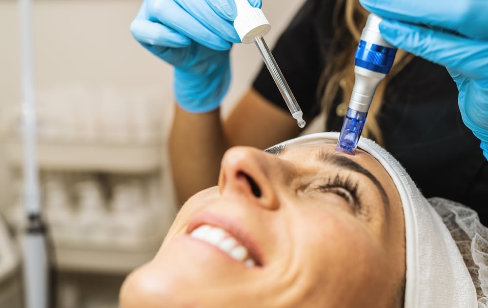Close-up of a persons face undergoing a skin treatment. A circular magnified area shows a mole being removed by a medical tool. The person has closed eyes, and the treatment focuses on a mole on the cheek.