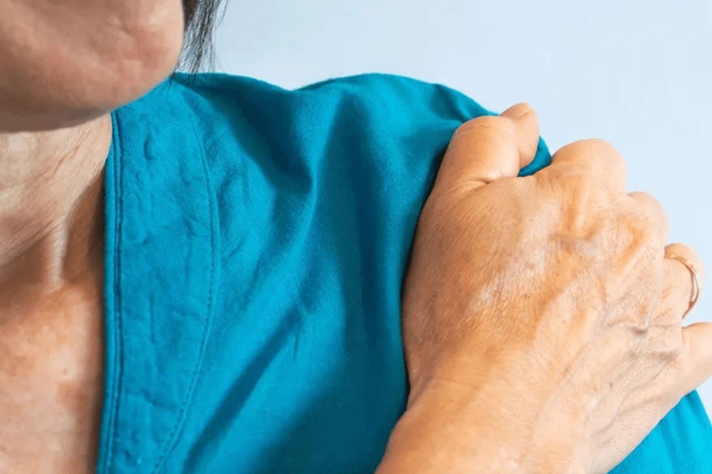 A healthcare professional in blue scrubs is taking the blood pressure of a woman sitting on a sofa. The woman is wearing a brown top and has short blonde hair. The setting is cozy, with a lamp and bookshelf in the background.
