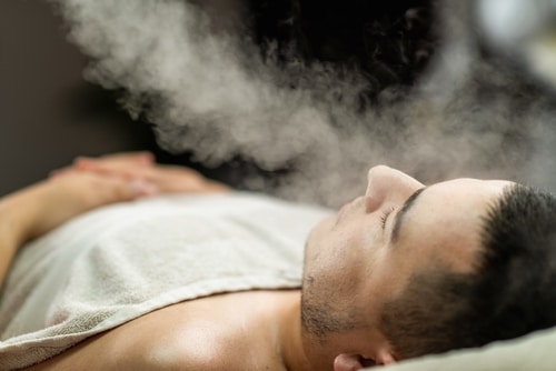 A person lying down with eyes closed receives a facial massage, wrapped in a towel, with hands gently massaging their cheeks and forehead.