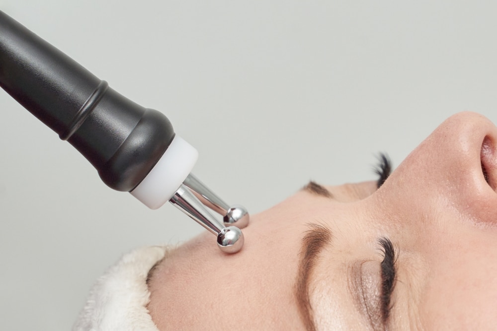 A doctor in a white coat examines a patients hand with a magnifying glass at a desk. The patient, with long brown hair, sits across from the doctor in a bright, sunlit room. Both are focused on the examination.