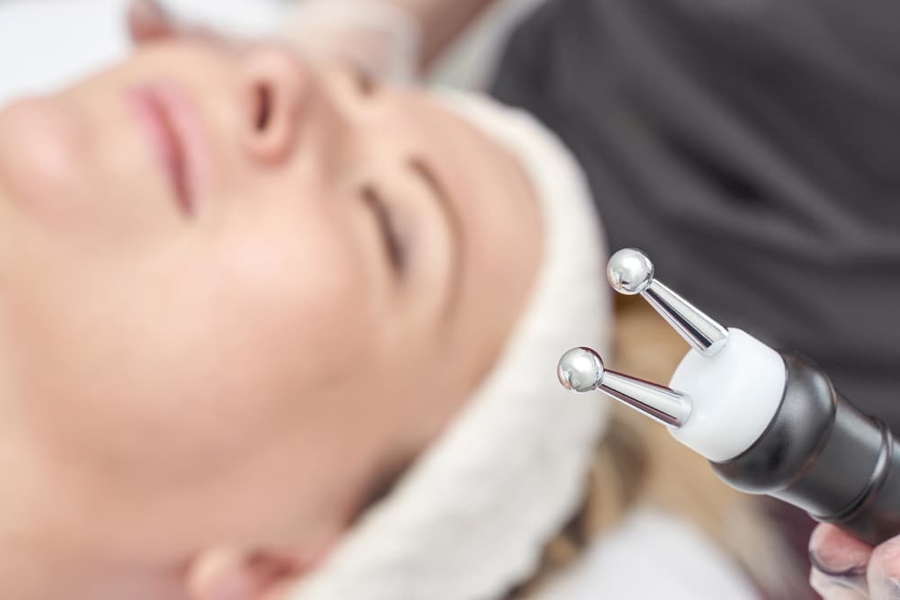 A doctor in a white coat examines a patients hand with a magnifying glass at a desk. The patient, with long brown hair, sits across from the doctor in a bright, sunlit room. Both are focused on the examination.
